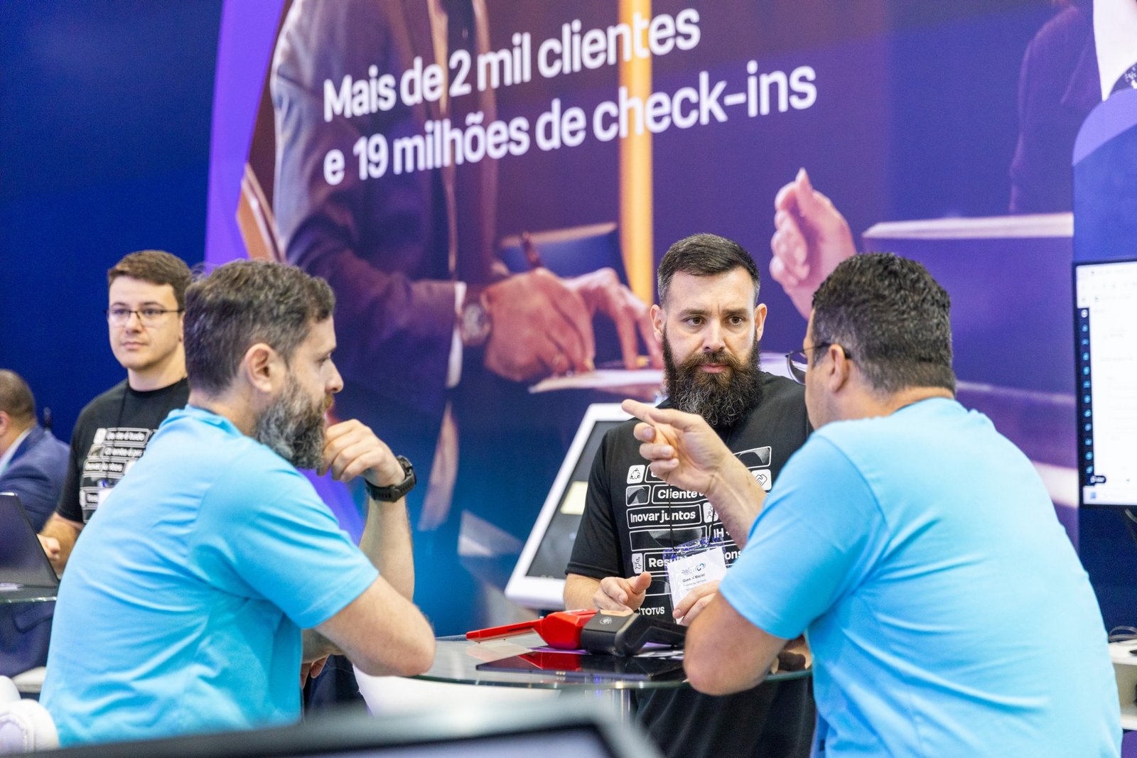 Três homens conversam em uma mesa durante rodada de negócios na Equipotel. Dois vestem camisetas azuis e o do centro usa camiseta preta. Ao fundo, um painel roxo exibe o texto "Mais de 2 mil clientes e 19 milhões de check-ins".
