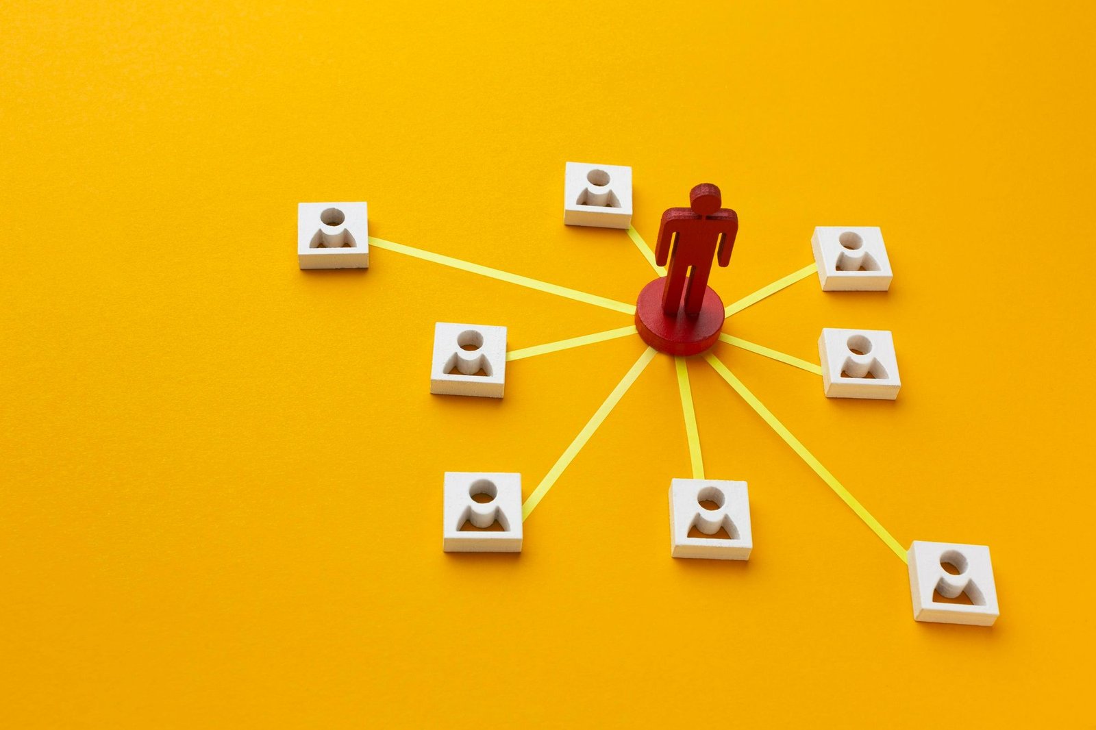 Vista superior de um diagrama feito com peças de madeira sobre fundo amarelo. Uma miniatura vermelha no centro conecta-se em rede a oito blocos brancos com ícones de colaboradores ao redor. A foto simboliza a integração de equipes e a visão sistêmica proporcionada pelo job rotation.