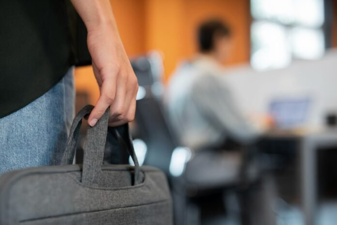 Imagem foca na mão de um funcionário segurando uma pasta de notebook cinza, como se ele tivesse acabado de chegar em um novo lugar. Ele veste calça jeans e camiseta preta. O fundo, desfocado, apresenta um escritório moderno em tons de laranja e outra pessoa trabalhando em frente a um computador. A foto simboliza o início de um novo ciclo e a transição entre áreas por meio do programa de job rotation.