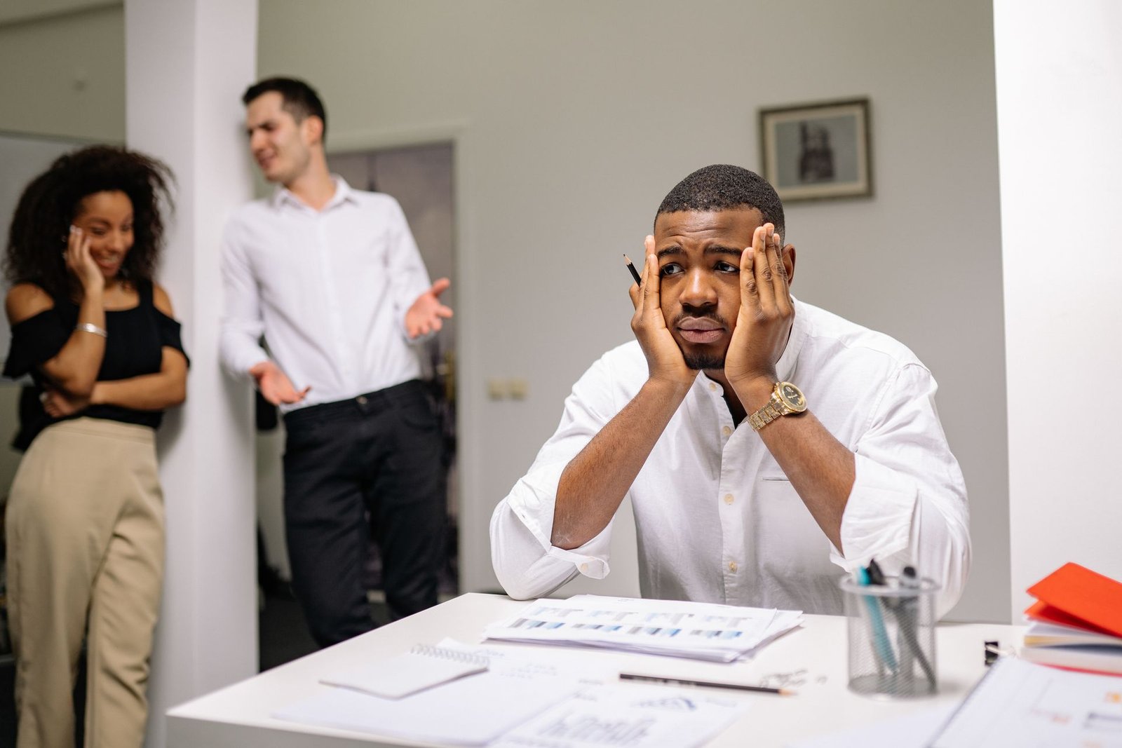 Homem negro em primeiro plano, sentado em uma mesa de escritório com papéis, apoia o rosto nas mãos com expressão de frustração e cansaço. Ao fundo, desfocados, dois colegas conversam e gesticulam. A imagem ilustra o estresse, a sobrecarga e o ruído de comunicação no ambiente de trabalho.