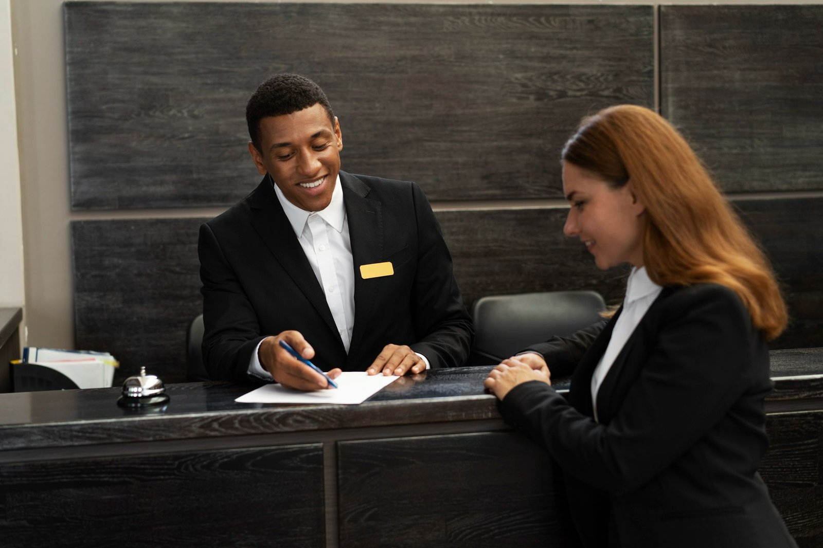 Recepcionista de hotel sorridente atende uma cliente no balcão da recepção, apontando com uma caneta para um documento sobre o balcão. A imagem contextualiza a necessidade de regras claras no room office para manter a eficiência e bom atendimento na recepção, prevenindo impactos negativos no fluxo de check-in e checkout.
