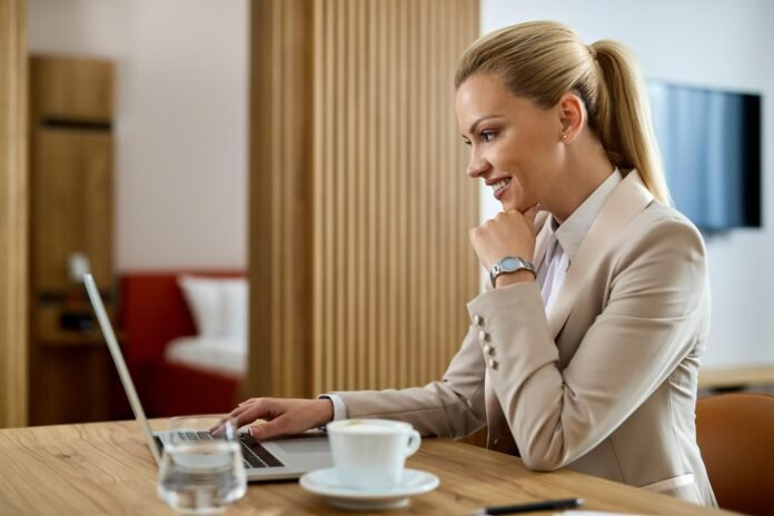 Mulher sorrindo enquanto trabalha no computador na mesa de um quarto de hotel. Imagem ilustra o uso do room office para otimizar a taxa de ocupação do hotel.
