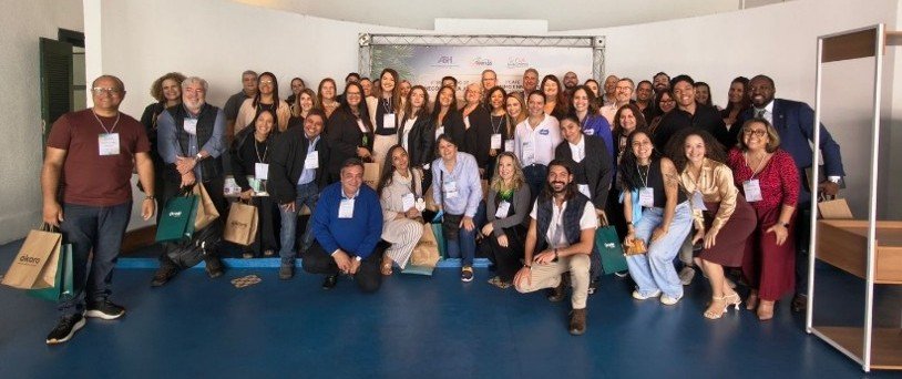 Uma fotografia de grupo de plano médio, tirada de um ângulo ligeiramente elevado, mostrando os participantes do Encontro de Negócios reunidos. O grupo diversificado de homens e mulheres está em pé e ajoelhado em várias fileiras, preenchendo todo o espaço. Todos estão sorrindo calorosamente e olhando para a câmera, transmitindo uma forte sensação de união e cooperação entre os membros do trade turístico do litoral de São Paulo.