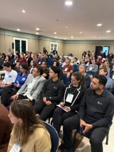 Vista em ângulo elevado de um auditório moderno, com teto rebaixado e iluminação embutida, completamente lotado. Centenas de participantes estão sentados em fileiras de poltronas azuis, todos voltados para a frente (palco não visível), atentamente ouvindo uma palestra durante o Encontro de Negócios. A diversidade de trajes de negócios e crachás de identificação reflete o público profissional. A composição captura a profundidade da sala preenchida por pessoas.
