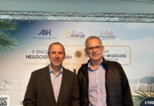 6º Encontro de Negócios do Portal do Hoteleiro e da ABIH-SP inova, firma parceria com o Visite Guarujá e reúne trade da Baixada Santista Marcos Vilas Boas, presidente da ABIH-SP, e o hoteleiro João Carlos Pollak sorriem lado a lado em pé. À esquerda, Marcos é um homem de pele clara, cabelos escuros e curtos, usando paletó escuro e camisa social azul sem gravata. À direita, João Carlos é um homem de pele clara, cabelos grisalhos curtos, usando óculos de grau e jaqueta preta sobre camisa azul. Ao fundo, um painel do 6 Encontro de Negócios exibe marcas da ABIH-SP, Visite Guarujá e do 1º Café, Turismo & Negócios.