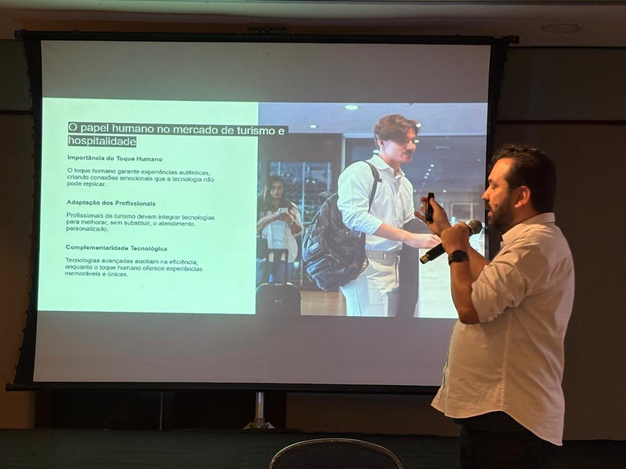 À direita da imagem, Fabio Zelenski, diretor de Marketing e Inovação do Visite São Paulo, ministra palestra em pé. Ele é um homem de pele clara, barba e cabelos escuros, vestindo camisa social clara com as mangas dobradas. Ele gesticula enquanto segura um microfone e um passador de slides. Ao fundo, uma grande tela de projeção exibe a apresentação com o título "O papel humano no mercado de turismo e hospitalidade", acompanhado de tópicos explicativos e a foto de um viajante em um hotel.