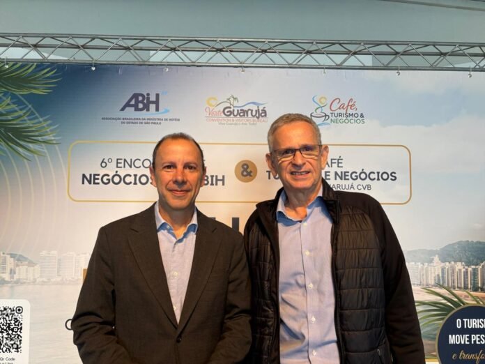 Marcos Vilas Boas, presidente da ABIH-SP, e o hoteleiro João Carlos Pollak sorriem lado a lado em pé. À esquerda, Marcos é um homem de pele clara, cabelos escuros e curtos, usando paletó escuro e camisa social azul sem gravata. À direita, João Carlos é um homem de pele clara, cabelos grisalhos curtos, usando óculos de grau e jaqueta preta sobre camisa azul. Ao fundo, um painel do 6 Encontro de Negócios exibe marcas da ABIH-SP, Visite Guarujá e do 1º Café, Turismo & Negócios.