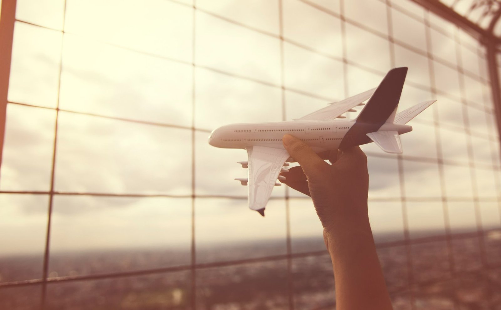Visão em close-up de uma mão segurando um modelo de avião de brinquedo perto de uma janela de aeroporto. A imagem é usada para ilustrar como a precificação dinâmica na aviação impacta outros setores turísticos, como a hotelaria.