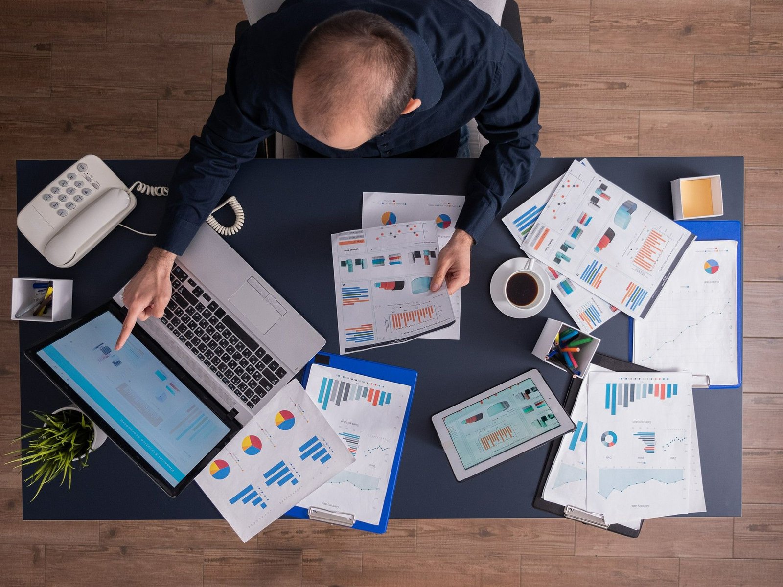 Visão superior de um homem em sua mesa de trabalho analisando dados de precificação dinâmica. Ele aponta para a tela do notebook cercado por um tablet, pranchetas com relatórios financeiros, gráficos coloridos e uma xícara de café.