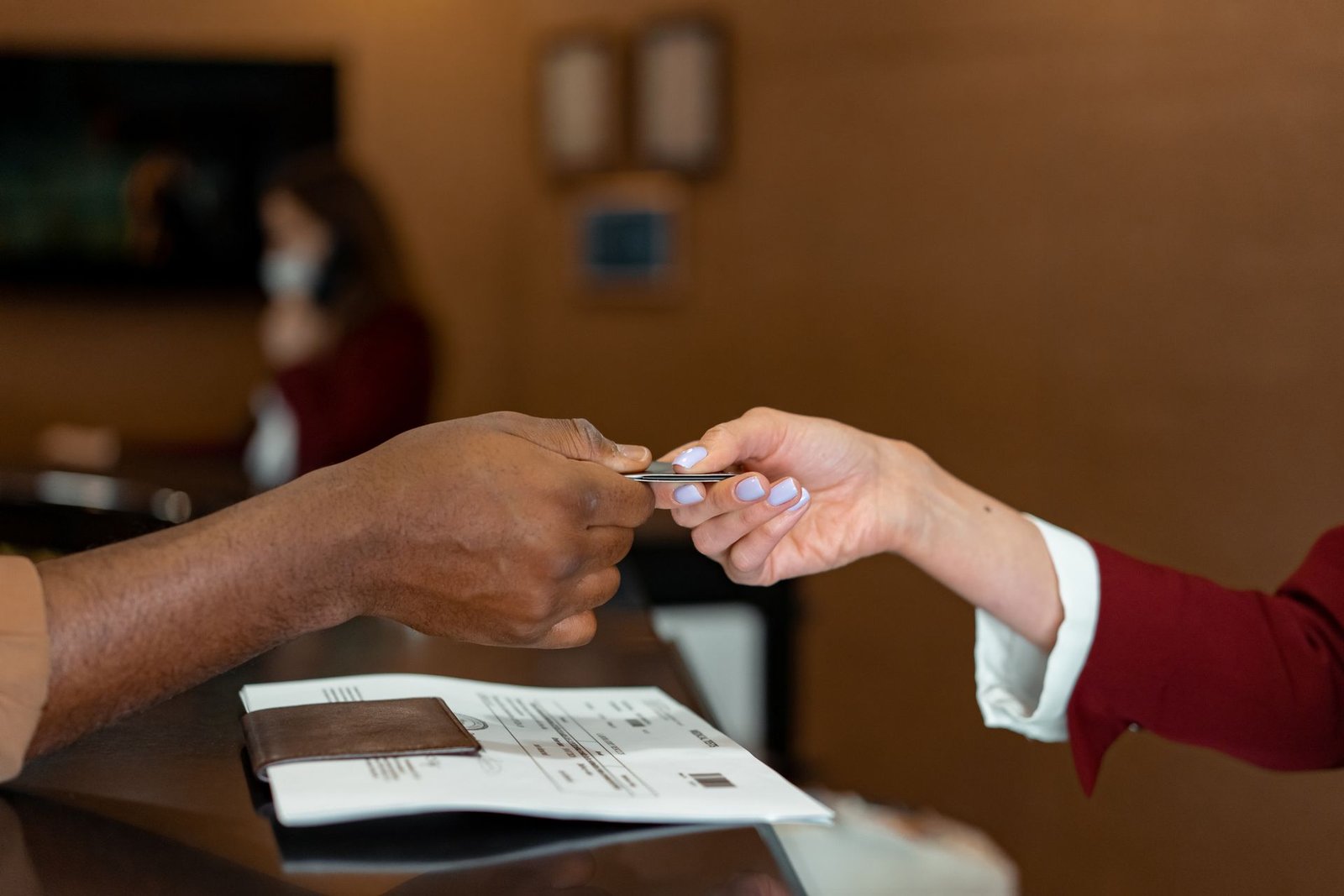 Hóspede recebe o cartão-chave do quarto das mãos da recepcionista no balcão de um hotel durante o check-in. Ao fundo, vê-se a recepção do estabelecimento.