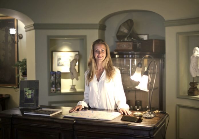 Mulher loira vestindo camisa branca sorri atrás de um balcão de madeira em uma recepção de pousada. Sobre o balcão há um mapa turístico, uma luminária clássica e uma sineta. A imagem simboliza o atendimento humanizado necessário à gestão de pousadas e hotéis pequenos.