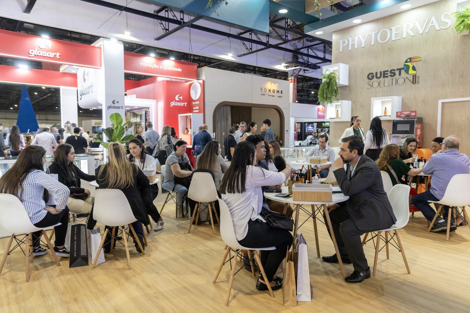 Profissionais do setor hoteleiro reunidos em pequenas mesas brancas durante as rodadas de negócios realizadas na Equipotel. Em primeiro plano e ao fundo, duplas e grupos conversam sentados em cadeiras brancas em um espaço de networking, com estandes de marcas como Glasart, SoHome e Phytoervas ao fundo do pavilhão de exposições.