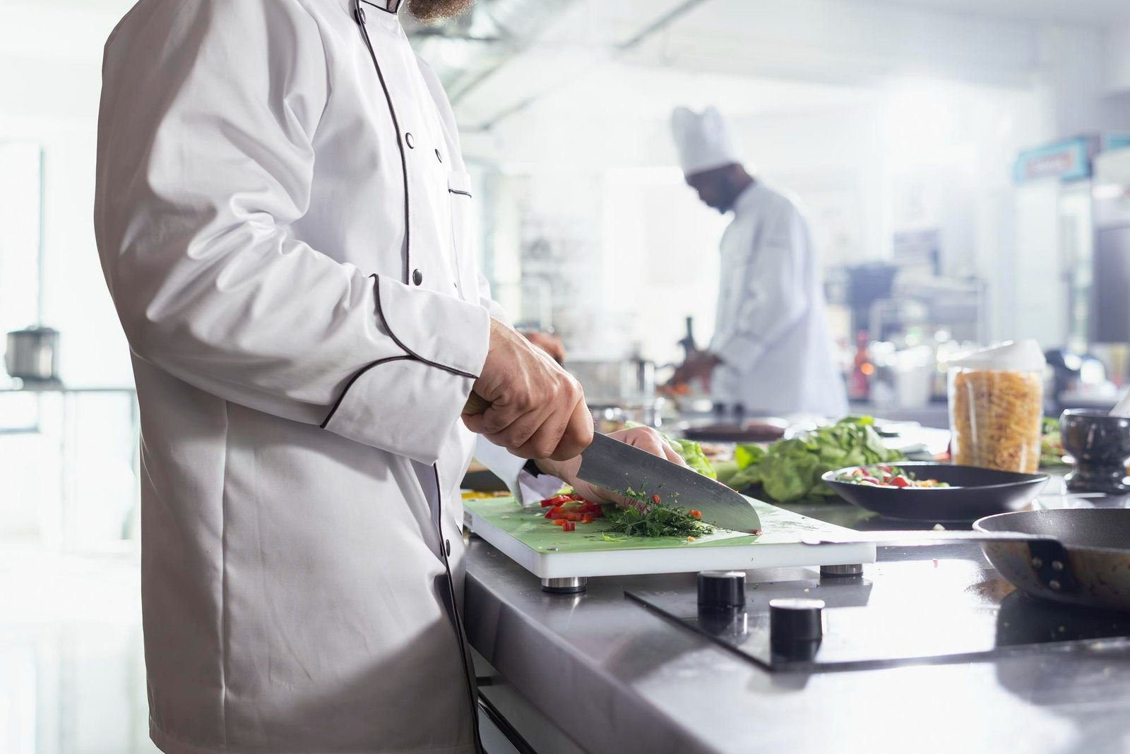 Cozinheiro em dólmã branco picando ervas e vegetais em uma tábua de corte em uma cozinha profissional iluminada, com outro chef trabalhando ao fundo. A imagem ilustra a preparação de alimentos e o alto volume de resíduos orgânicos gerados diariamente pelos times de A&B dos hotéis.
