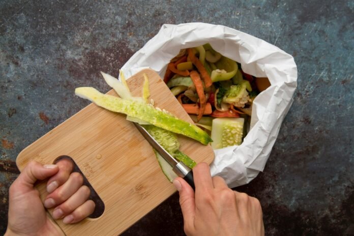 Vista superior de mãos raspando cascas de vegetais de uma tábua de madeira para dentro de um saco de papel com restos orgânicos, exemplificando a prática de gestão de resíduos na cozinha — a qual pode ser aperfeiçoada com o uso de tecnologia.