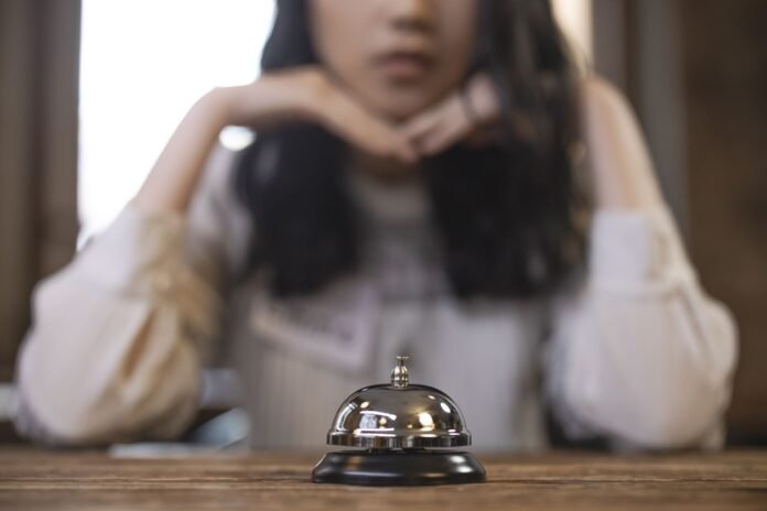 Sino prateado de recepção em foco sobre um balcão de madeira. Ao fundo e desfocada, uma recepcionista apoia o queixo nas mãos, demonstrando cansaço e apreensão e transmitindo o sentimento de ansiedade no trabalho.