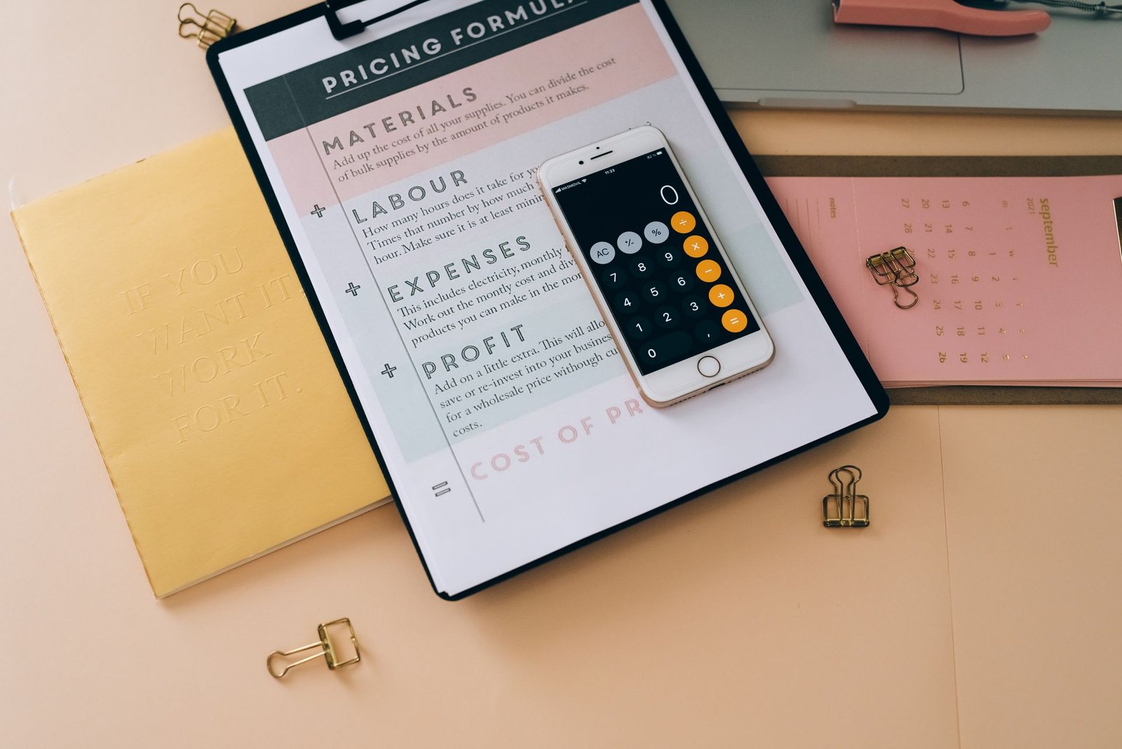 Smartphone com calculadora aberta sobre prancheta de fórmula de precificação, exibindo custos, despesas e margem de lucro.