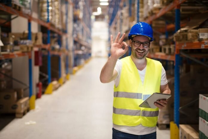 Um trabalhador de armazém sorridente, com capacete azul, colete de alta visibilidade amarelo e segurando um tablet, faz um sinal de 