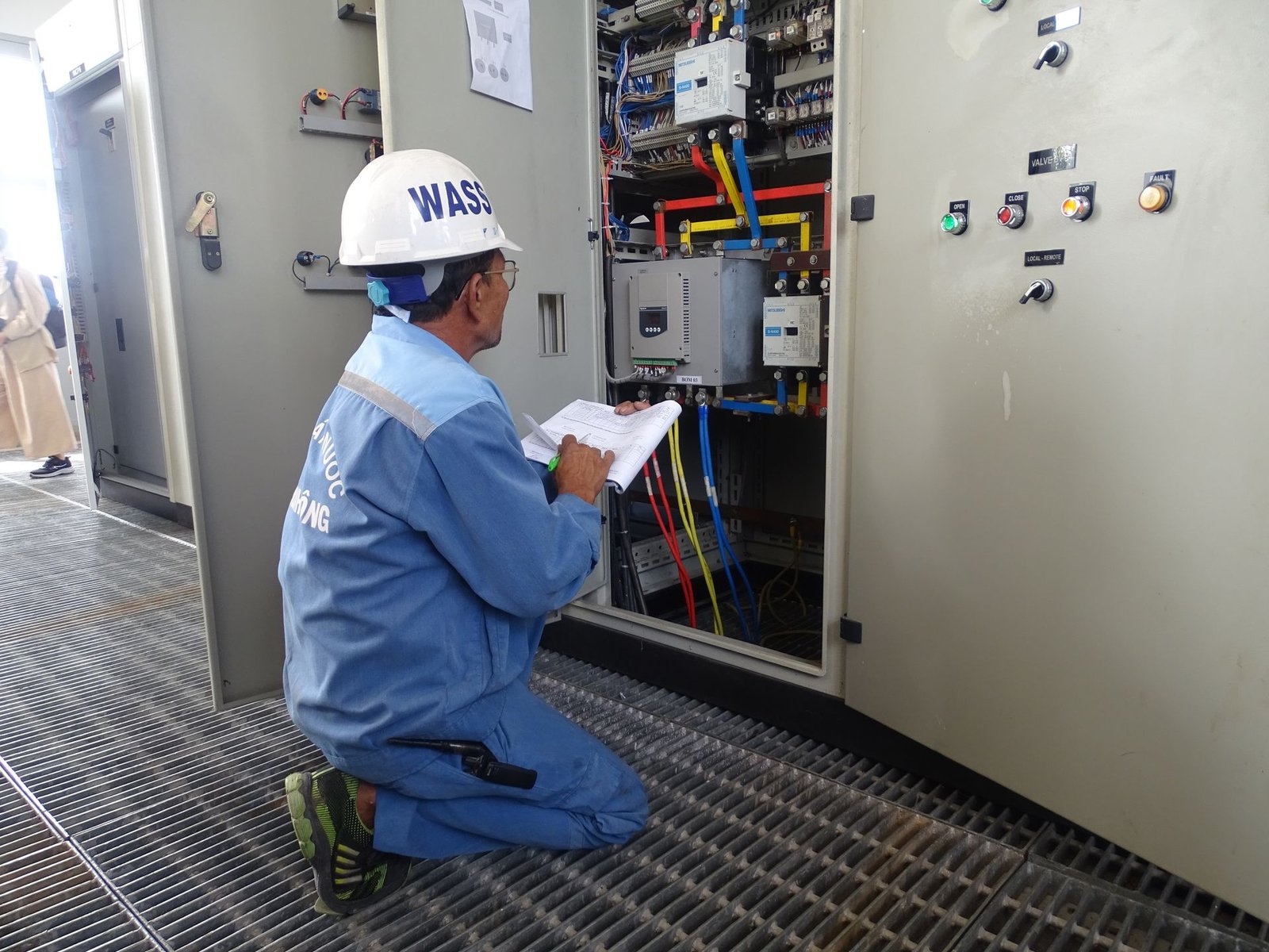Fotografia de plano médio de um técnico de manutenção de joelhos em uma casa de máquinas de hotel. Ele veste uniforme azul de segurança, capacete branco e examina com atenção um painel elétrico central aberto, repleto de fiações e componentes. Ele segura um bloco de anotações na mão para registrar dados de medição. A imagem ilustra o monitoramento prático executado pela equipe de manutenção para garantir indicadores auditáveis de ESG.