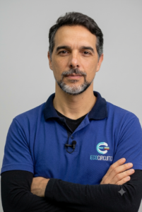 Retrato de Eduardo Prates, fundador da Eco Circuito, um homem de cabelos escuros grisalhos e barba aparada. Ele está de braços cruzados, olhando diretamente para a câmera com uma expressão neutra, vestindo uma camisa polo azul-marinho com o logotipo da Eco Circuito bordado no peito e uma blusa preta de mangas compridas por baixo. O fundo é neutro e em tom cinza-claro.