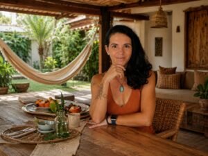 Nathalia Borghi, criadora do projeto Viver de Pousada, posa sorrindo em uma varanda rústica de pousada. Ela está sentada à frente de uma mesa de madeira posta para o café da manhã, com o queixo apoiado na mão. Ao fundo, vê-se um jardim tropical com uma rede de descanso, representando hospitalidade e natureza.