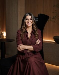 Renata Freire, criadora da empresa Mkt dê Sentidos, posa sorrindo com os braços cruzados sentada em uma poltrona preta. Ela veste um vestido de manga longa na cor vinho e está em um ambiente moderno e aconchegante, com iluminação quente.