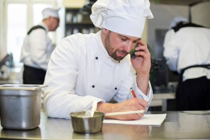 Chef de cozinha vestindo dólmã e chapéu brancos fala ao telefone enquanto faz anotações em um papel, ilustrando a gestão de alimentos e bebidas na hotelaria. Ao fundo, outros profissionais uniformizados aparecem trabalhando.