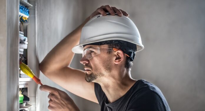 Técnico de manutenção usando capacete e óculos de proteção enquanto segura uma chave de fenda e inspeciona um quadro elétrico aberto, representando a complexidade relacionada à gestão da infraestrutura hoteleira.