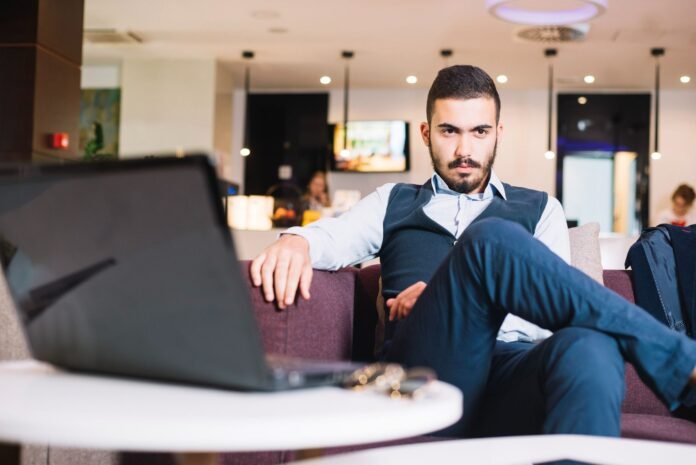 Homem de camisa social e colete com expressão pensativa, sentado em um sofá no lounge de um hotel. À sua frente há um notebook aberto. A imagem representa a complexidade do trabalho de marketing hoteleiro e a reflexão inerente à análise de dados.