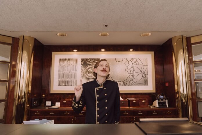 Funcionário com uniforme clássico e bigode estilizado posicionado atrás de uma recepção com painéis de madeira, fazendo um gesto de atenção com o dedo erguido, como se quisesse chamar a atenção do RH na hotelaria.