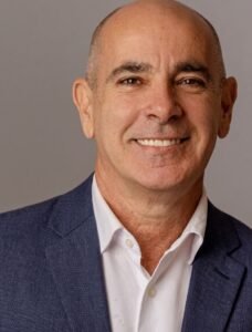 Foto em enquadramento fechado (plano médio) de Heber Garrido, executivo da ABIH-SP. Ele é um homem de pele clara, calvo, com olhos castanhos, sorrindo para a câmera. Veste um paletó azul-marinho sobre uma camisa social branca de colarinho aberto. O fundo é neutro, em tom claro de cinza.